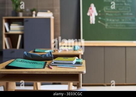 Empty classroom showing wooden desks with health education folders, notebooks, and pencils arranged neatly, green chalkboard with human anatomy diagram visible in background Stock Photo