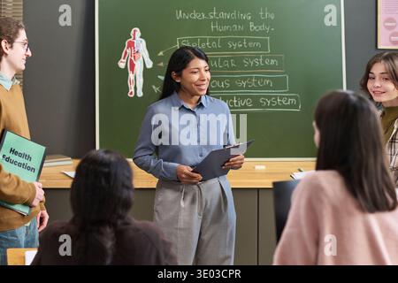 Hispanic woman teacher explaining human body systems to group of teenagers in classroom, holding clipboard, students listening attentively Stock Photo