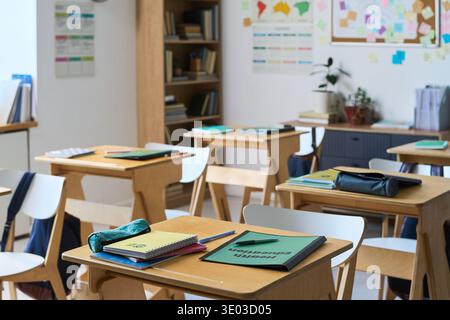 Empty classroom showing wooden desks with notebooks, folders, and stationery scattered on surfaces, bookshelves filled with books in background, educational posters and bulletin board on wall Stock Photo