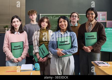 Group of multiethnic teenagers standing in classroom holding Health Education folders, smiling and facing camera, chalkboard with health topics in background, diverse gender representation Stock Photo