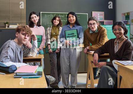 Group of diverse teenagers and young adult female teacher standing and sitting in classroom holding health education materials, smiling and looking at camera during lesson Stock Photo