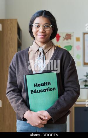 Vertical portrait of young adult Black woman smiling and holding health education folder, standing in classroom setting with educational materials visible in background, wearing glasses Stock Photo
