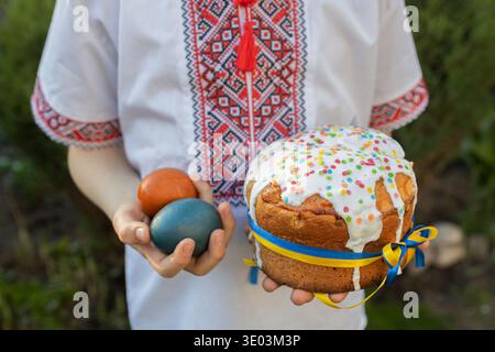 child in an embroidered shirt holds an Easter cake tied with a yellow and blue ribbon in one hand and painted eggs in the other. Easter theme. Ukraini Stock Photo