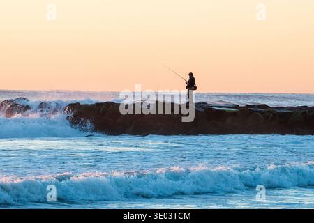 Silhouette of a fisherman standing on a rocky jetty as waves crash dramatically against it at sunrise, casting a warm orange glow across the sky Stock Photo