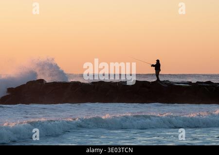 Silhouette of a fisherman standing on a rocky jetty as waves crash dramatically against it at sunrise, casting a warm orange glow across the sky Stock Photo