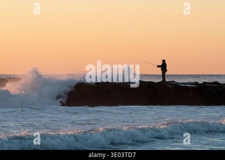 Silhouette of a fisherman standing on a rocky jetty as waves crash dramatically against it at sunrise, casting a warm orange glow across the sky Stock Photo