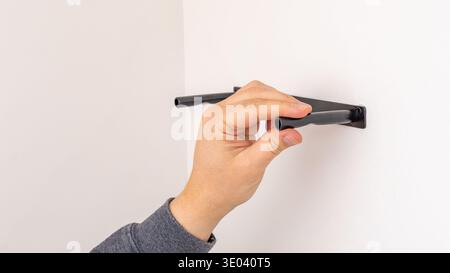 Mans hand using black marker to mark line on white wall above black shelf bracket. Home improvement, measuring and marking, DIY level indication, prec Stock Photo
