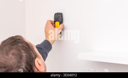 Mans hand using yellow cordless drill to fasten screw into white wall near black shelf bracket. Home improvement, additional securing, DIY reinforceme Stock Photo