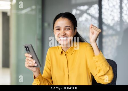 Happy young multiracial woman celebrating a successful online achievement on her smartphone, smiling brightly and raising a fist in triumph at the modern office Stock Photo