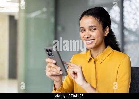 Young woman smiling at camera, holding and scrolling through a mobile phone, representing remote communication and technology in a modern office setup Stock Photo