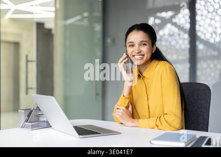 Young businesswoman with a cheerful expression talking on a cellphone, sitting at a desk with a laptop while working in a contemporary office building Stock Photo