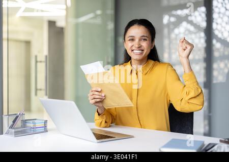 Happy young multiracial business woman celebrating achievement, expressing excitement while holding an important letter in an envelope at her modern office desk with a laptop Stock Photo
