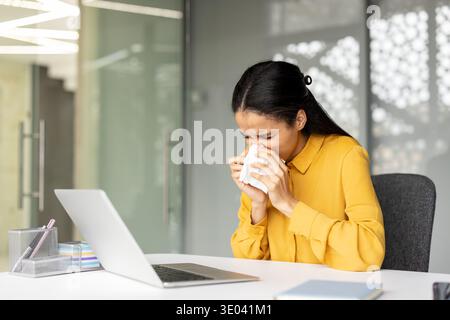 Young biracial woman sitting at her office desk, blowing her nose into a tissue, suffering from a common cold, flu, virus, or seasonal allergy symptoms while working Stock Photo
