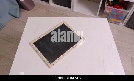 Small Blackboard Resting On White Surface. Compact Chalkboard With Aged Wooden Frame On Plain Table. Little Chalkboard With Worn Frame And Faint Chalk Stock Photo