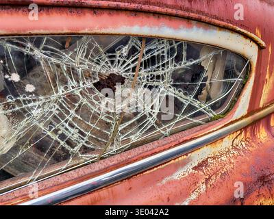 A spiderweb of cracks spreads through a shattered window on an old, rusting car in a junkyard. Stock Photo