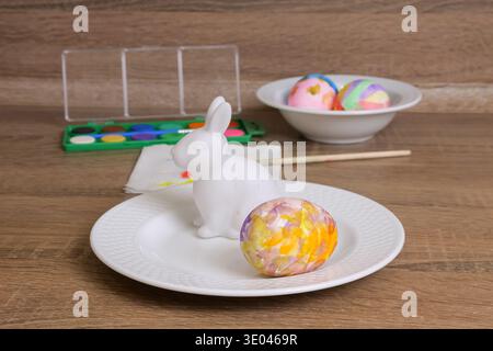 Closeup Of Bunny Figurine On Plate With Marbled Egg, Watercolor Palette, Candles, Bowl Of Decorated Eggs On Wood Table Warm Cozy Spring Craft Mood Stock Photo