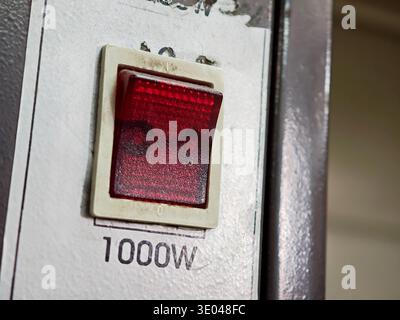 Old heavy-duty red illuminated rocker power switch labeled 1000W on a worn metal device panel, closeup of an industrial appliance on off control butto Stock Photo