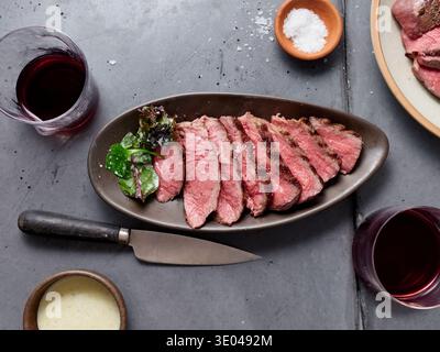 A beautifully sliced steak is served on a dark plate, accompanied by fresh greens and a sprinkle of seasoning. A glass of red wine adds an elegant tou Stock Photo