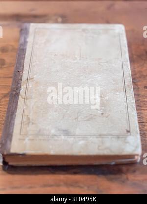 An aged book with a textured, weathered cover lies on a rustic wooden table. The hardcover features subtle embossed details, hinting at its long histo Stock Photo