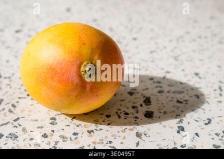 A ripe, colorful mango sits on a speckled terrazzo surface, showcasing its vibrant yellow and red hues under natural lighting. The detailed texture hi Stock Photo