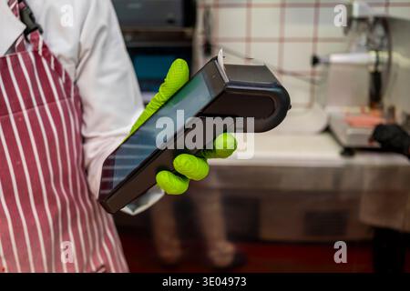 A butcher in a striped apron and green gloves uses a handheld card reader. Fresh meat and busy work environment are visible in the background, emphasi Stock Photo