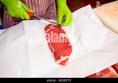 A butcher in a striped apron and green gloves carefully slices a piece of fresh beef on white paper. The work showcases precision and skill in meat pr Stock Photo