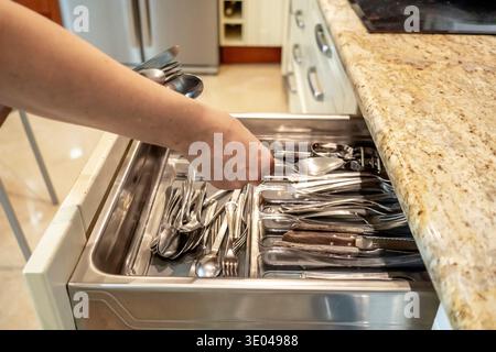 A hand reaches into an open kitchen drawer filled with neatly organized cutlery, including spoons, forks, and knives, in a modern kitchen setting with Stock Photo