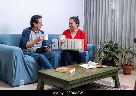 Mature couple sitting on sofa using digital devices and drinking coffee together Stock Photo