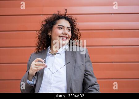 A confident woman with curly hair and glasses, dressed in a professional suit, stands against a modern orange backdrop. Her smile reflects positivity Stock Photo