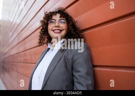 A confident woman with curly hair and glasses smiles broadly while leaning against a red wall. She is dressed in a formal blazer over a white shirt, e Stock Photo