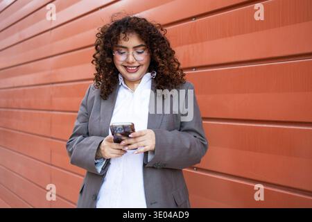 A businesswoman with curly hair, wearing a gray suit, smiles while using her smartphone. She stands against an orange wall, appearing confident and fo Stock Photo