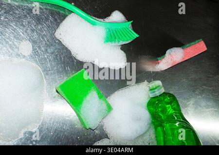 A collection of cleaning tools, including a green brush and sponges, is displayed on a metal surface covered in soap and foam, showcasing a domestic c Stock Photo