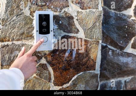 Hand Pressing Video Doorbell on Stone Wall. Close-up of a hand pressing a video doorbell on a stone wall, illustrating smart home entry, access Stock Photo