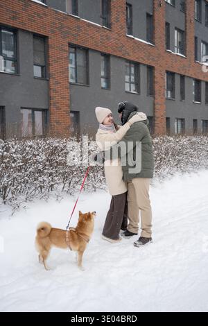 A joyful couple embraces during a winter walk, while their dog watches them excitedly, enjoying the snowy path. Snowflakes gently fall against the bac Stock Photo