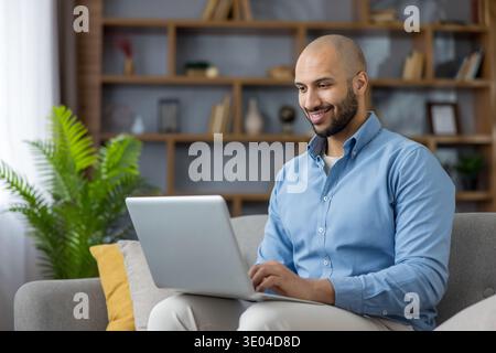 Young bald man smiling, using a laptop while sitting on a comfortable couch in a living room, happily engaging in remote work and managing online task Stock Photo