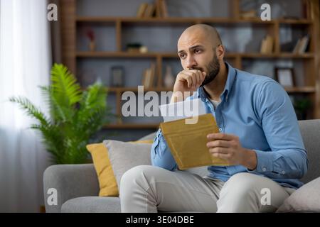 Man feeling anxious and stressed while holding an open envelope with a letter, contemplating financial problems, debt, or an unemployment notice, sitt Stock Photo