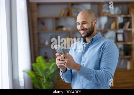 Smiling bearded bald man in a blue shirt using his smartphone indoors, happily texting and browsing, illustrating modern digital communication, connec Stock Photo