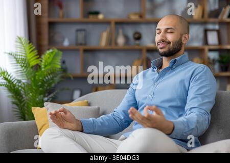 Young man seated on a living room couch with eyes closed, practicing mindful meditation for calm, inner peace and relaxed well-being in a cozy modern Stock Photo