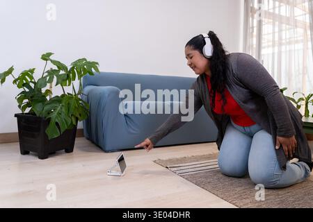 Woman on knees wearing headphones interacting with smartphone in living room Stock Photo