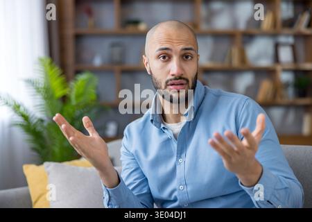 Young man sitting on a sofa, looking at the camera with a puzzled expression and gesturing with open hands, conveying doubts and seeking an explanatio Stock Photo