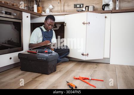 Plumber writing on clipboard while inspecting pipes under kitchen sink Stock Photo