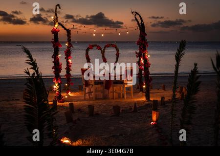 A romantic dinner table for two set on a beach at sunset with flower arches and decorative lights. Location: Zanzibar, Tanzania. Stock Photo