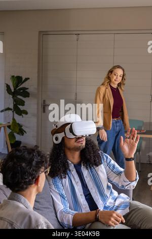 Diverse friends watching man using VR headset on gray sofa in lounge, woman in tan blazer Stock Photo