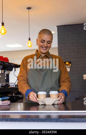 Female barista wearing green apron smiling placing two takeaway cups in cup carrier on cafe counter Stock Photo