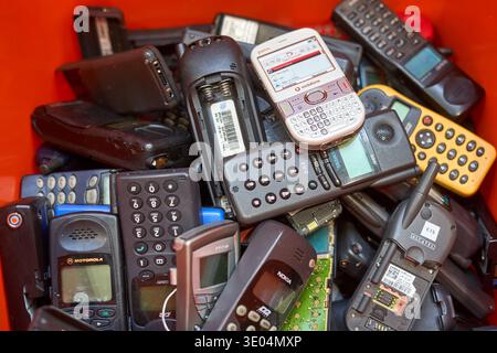 A close-up view of a pile of obsolete mobile phones and electronic devices stacked together in a bright orange container. The collection includes... Stock Photo