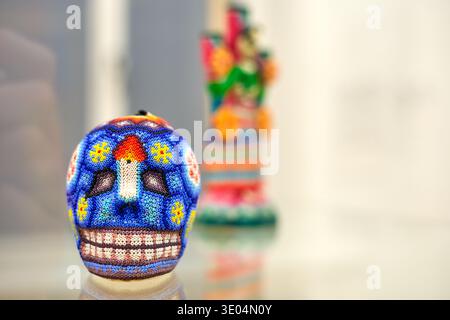 A vibrant, intricately decorated Mexican sugar skull (calavera) covered in colorful beadwork sits in sharp focus on a reflective glass surface. The.. Stock Photo