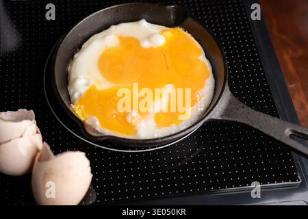 close-up of two eggs frying in a small cast-iron skillet on a black glass-ceramic stovetop with cracked eggshells nearby; warm golden yolks and glossy Stock Photo