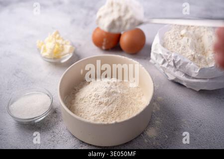 close-up of a ceramic bowl filled with flour surrounded by eggs, butter, sugar and an open paper flour bag on a textured gray countertop, soft natural Stock Photo