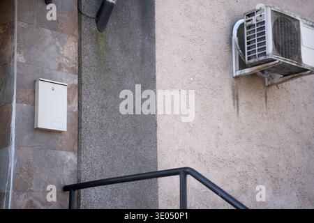 urban corner showing white mailbox on tiled cladding, textured concrete and stucco walls, an aged wall air-conditioner, black metal handrail and drain Stock Photo
