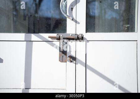 sunlit white metal double door with a weathered rusty sliding bolt lock and twisted latch, chrome curved handle, dusty upper glass panes reflecting bl Stock Photo